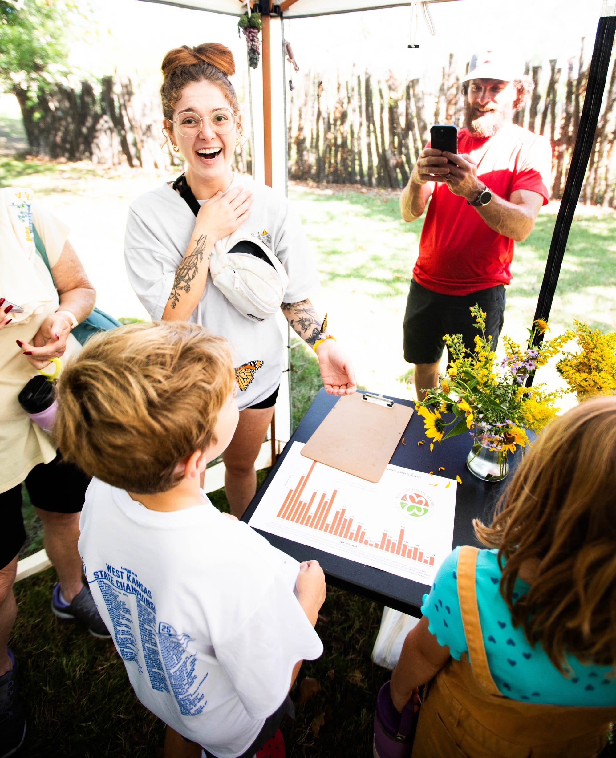A woman, hand on her heart, smiles in disbelief with a monarch butterfly on her arm while a man takes a photo of monarch butterfly perched on a child's face
