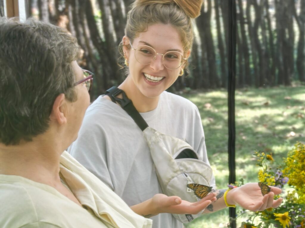 A smiling guest shows off a monarch butterfly in each hand to another guest.