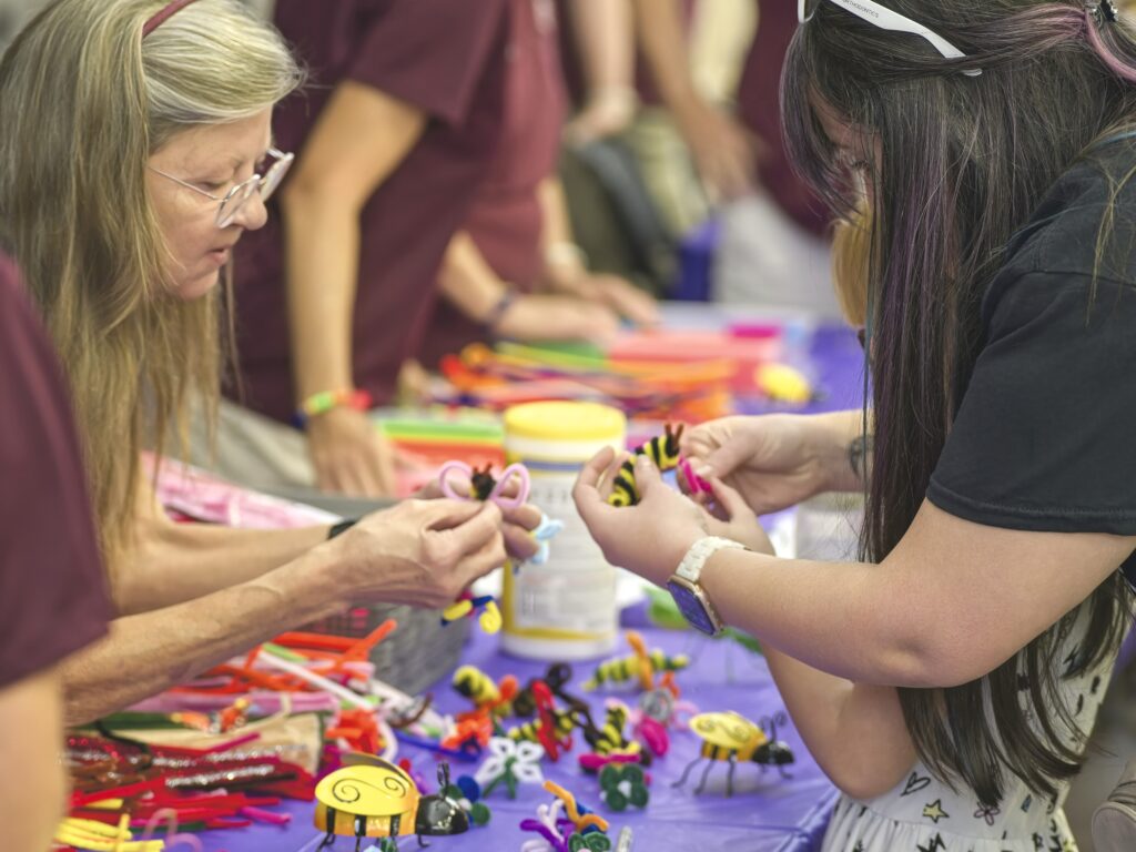A Master Gardener volunteers leans forward over a table filled with beads and other craft supplies to help a child build a pollinator bracelet