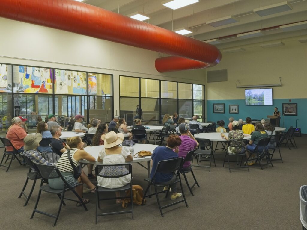 A room of table and chairs full of people arranged in front of a projector screen.